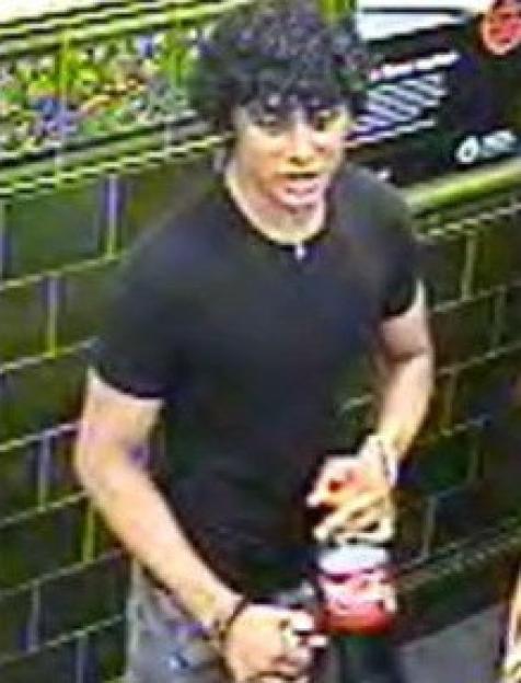 A young man with curly dark hair wearing a black t-shirt and holding a red bottle.
