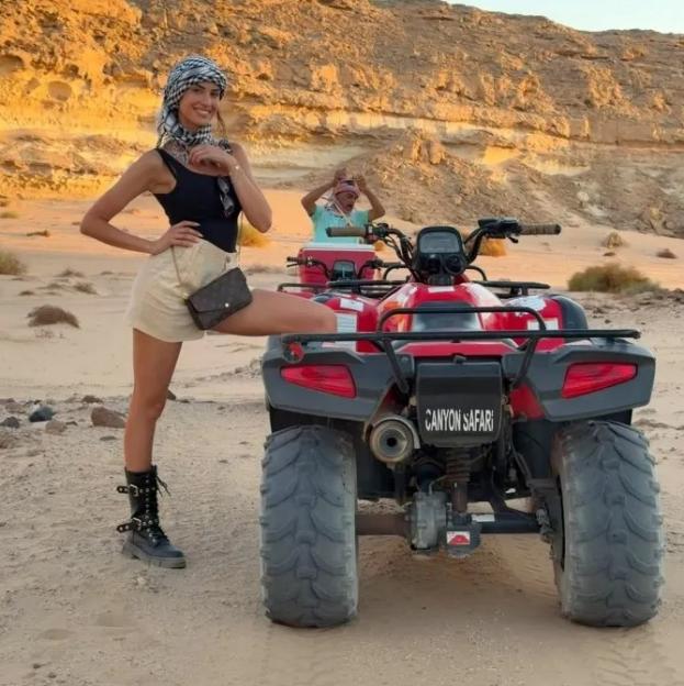 A woman in a keffiyeh and a man in the background next to red ATVs in a desert landscape.