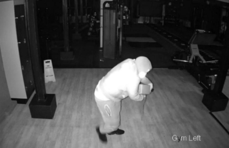 Security footage of a person in a hooded sweatshirt carrying a brick through a gym at night.