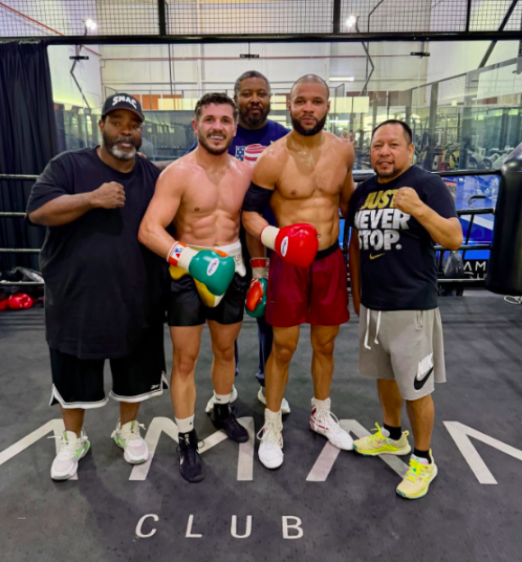 Five men in a boxing gym, two in boxing attire with gloves, and three in casual clothing.