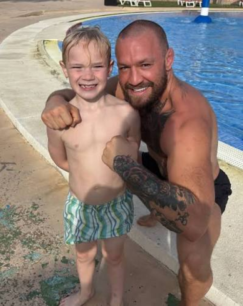 Conor McGregor flexing his biceps next to a young boy at a swimming pool.