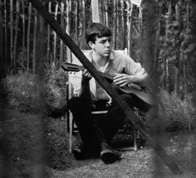 Paul McCartney sitting on a chair in a backyard holding a guitar, viewed through a window with clothes on a line.