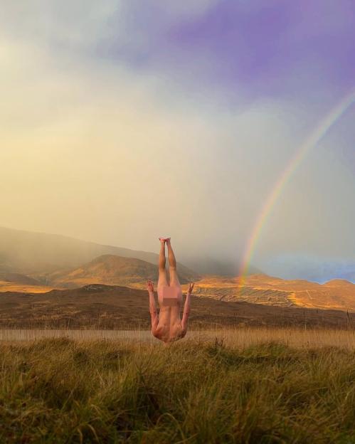 A person in a handstand with pixelated genitalia in a field with a rainbow and mountains in the background.