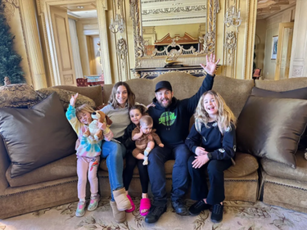 A family of six, including four children of various ages, sit on a couch in a lavish room, smiling and waving.