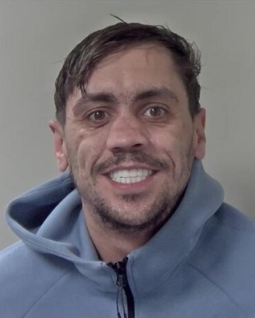 A man with short brown hair, stubble, and a light blue hoodie smiles at the camera.