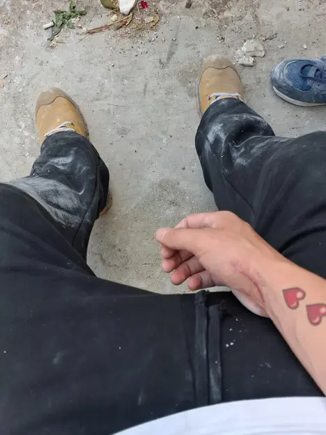 A person's legs and right arm with a scar and heart tattoos, wearing dirty black pants and work boots, seen from a first-person perspective.