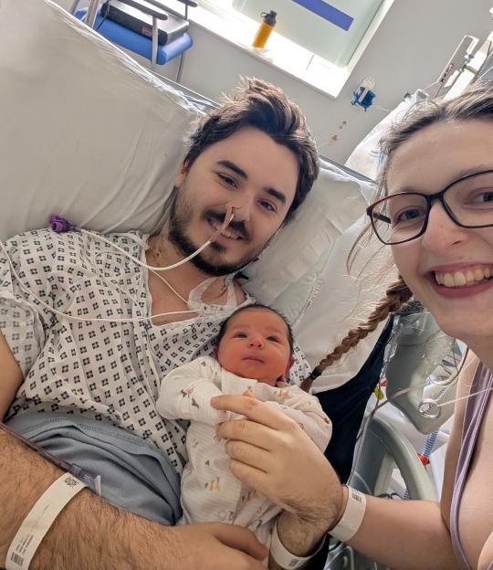 A man lying in a hospital bed with a baby on his chest, while a woman wearing glasses smiles at the camera, taking a selfie.