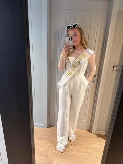 Jess-Hulley Jones taking a mirror selfie wearing a white jumpsuit and a "Bride-to-be" sash.