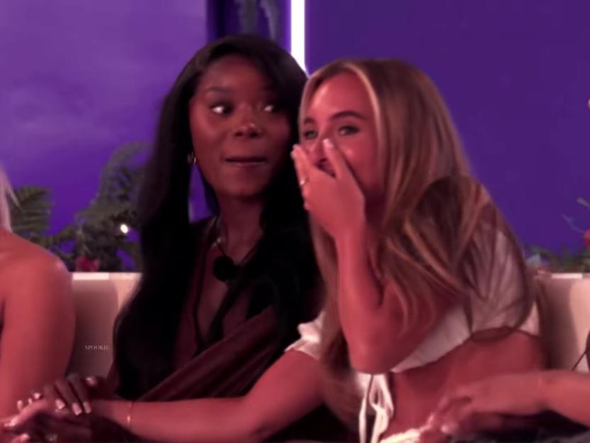 A dark-skinned woman with dark hair looks on as a light-skinned woman with blonde hair covers her mouth in surprise.