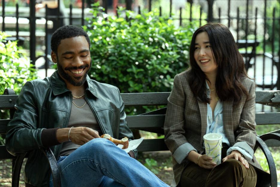 Donald Glover and Maya Erskine sitting on a park bench, smiling.