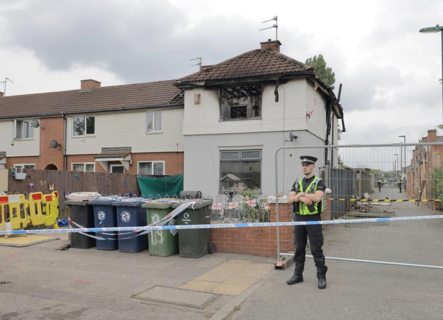 A police officer stands guard at a fire-damaged house, cordoned off with police tape.