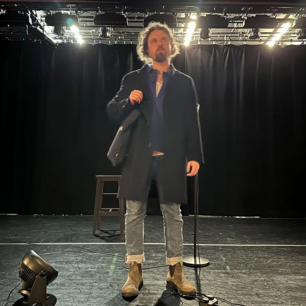 Alfie Brown standing on a stage with a microphone stand, chair, and floor light.