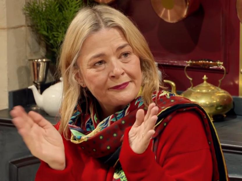 A woman with blonde hair, a red shirt, and a colorful scarf looking up with her hands raised.