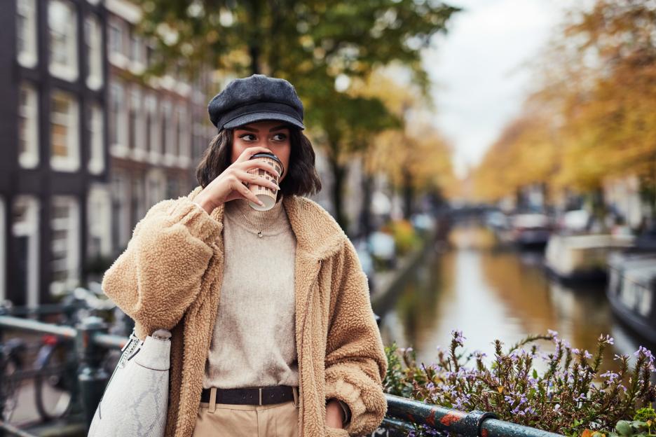 A young woman in a fuzzy coat and cap sips coffee by a canal in the city.