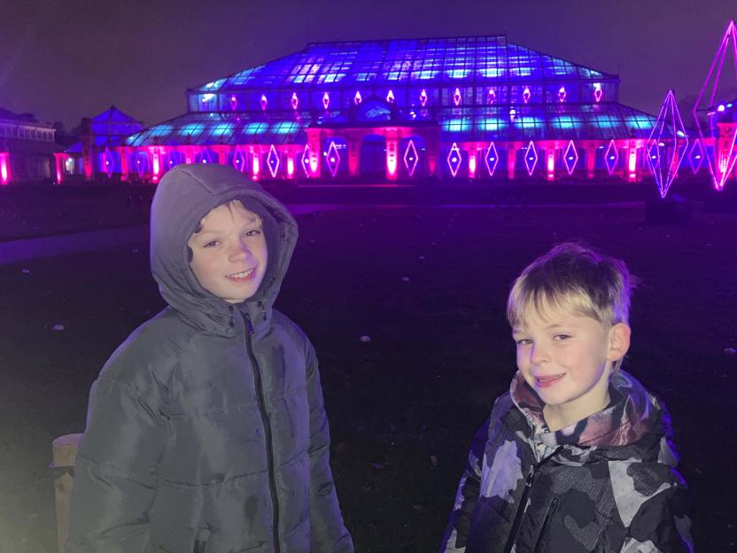 Two young boys smile in front of the Kew Gardens light trail with purple and blue lights.