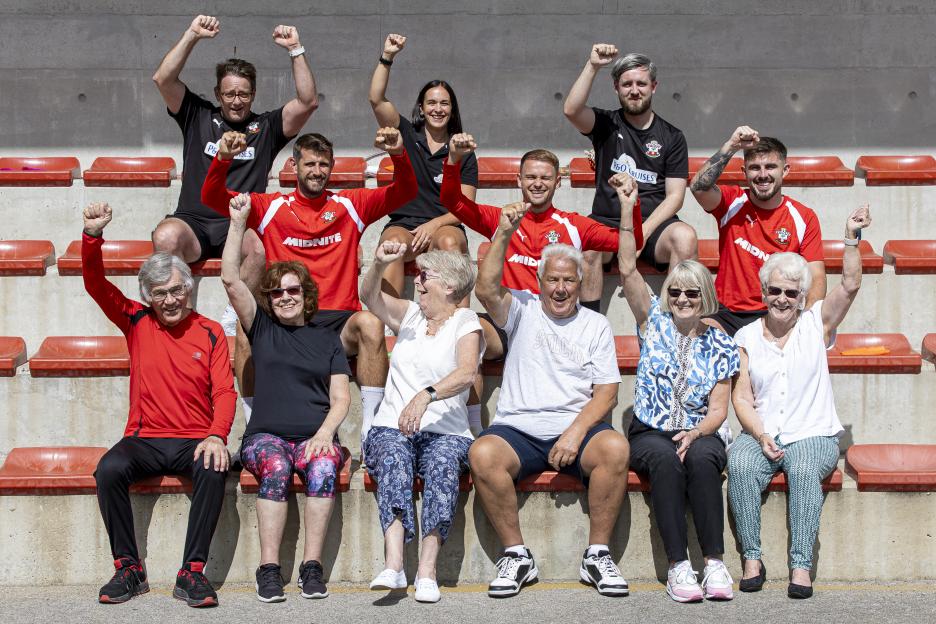 A group of older people and younger adults, all wearing athletic apparel, sitting in bleachers with their fists raised in the air.