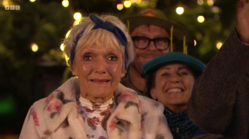 An elderly woman with a blue headscarf and a floral jacket smiles in the foreground, with two other people partially visible behind her.