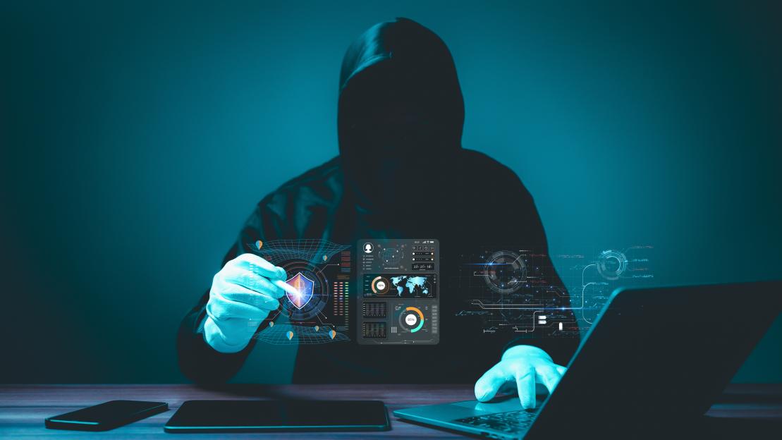 A person in a hooded sweatshirt and gloves works on a laptop, with a holographic shield icon and data analysis graphics floating in front of them, symbolizing cybersecurity.