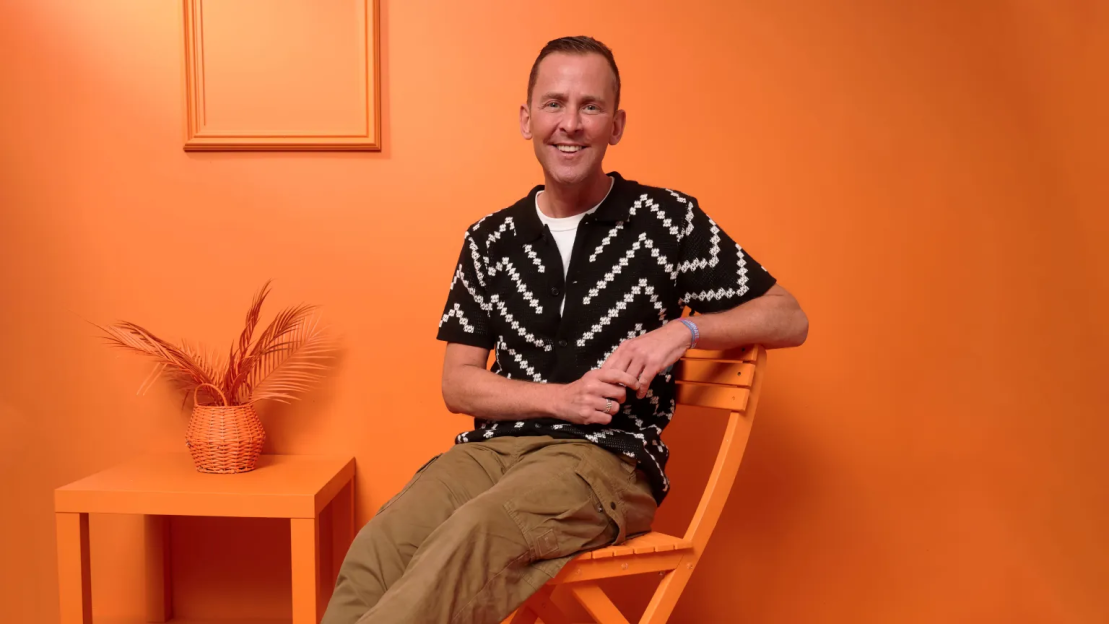 A man with short brown hair, wearing a patterned short-sleeved shirt and cargo pants, sitting on an orange chair in a monochromatic orange room.