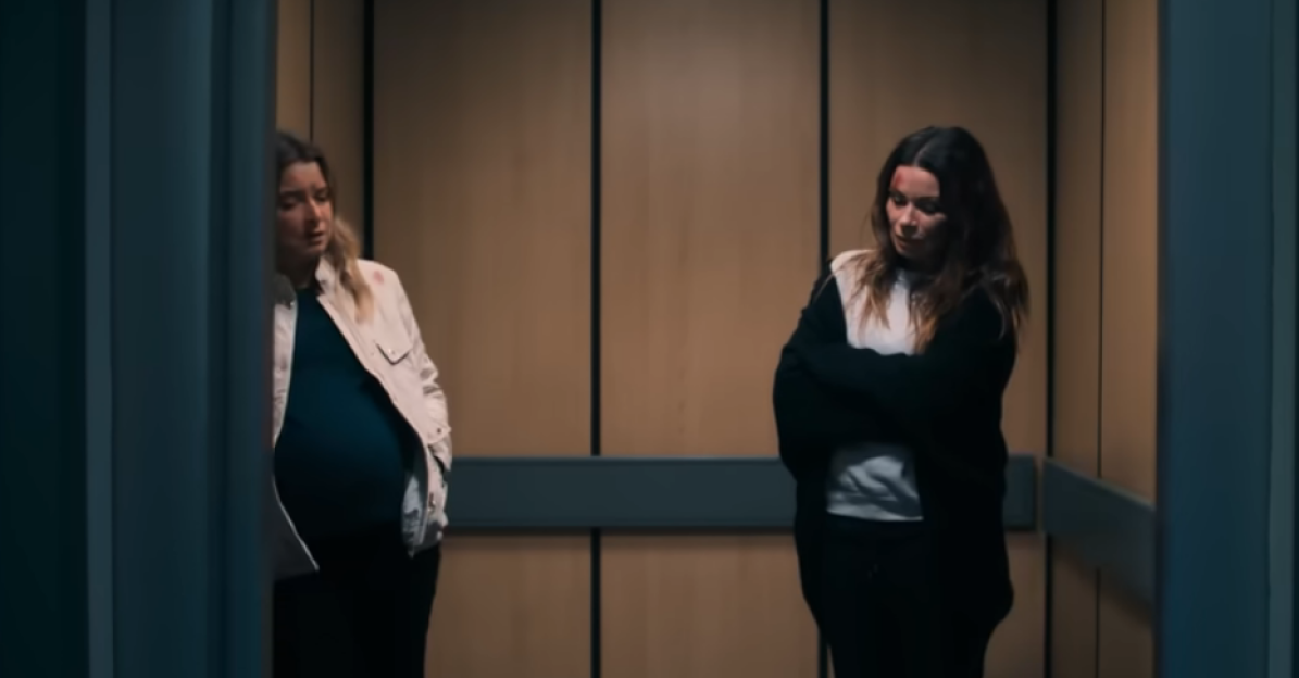 Two women in an elevator, one visibly pregnant, both looking downcast.