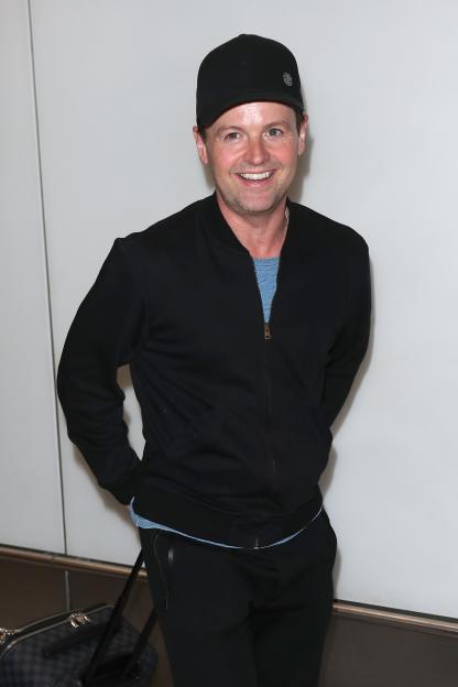 Declan Donnelly smiling and wearing a black jacket and cap at Heathrow Airport.
