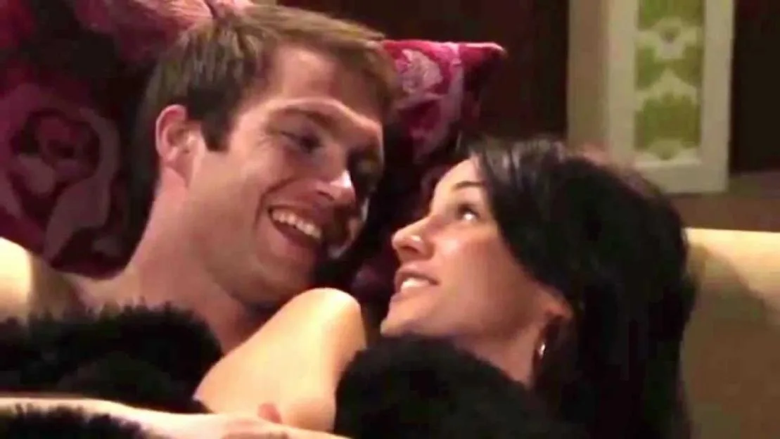 A shirtless man laughing and a woman smiling at him from bed.