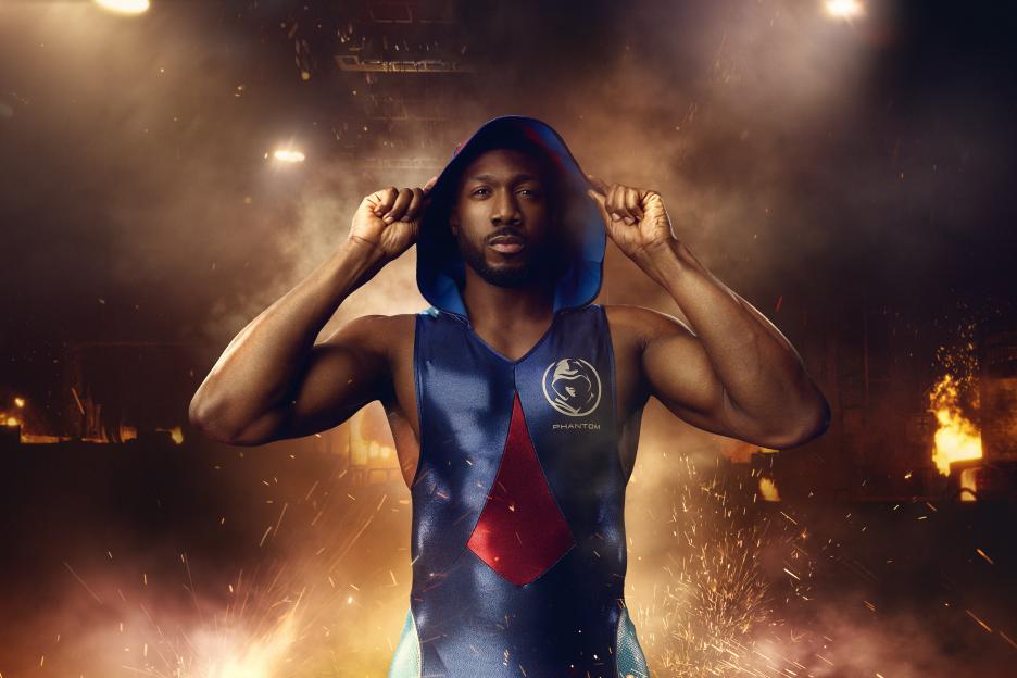 Phantom, a male Gladiator, poses in a blue and red outfit with his hood up against a background of sparks and smoke.