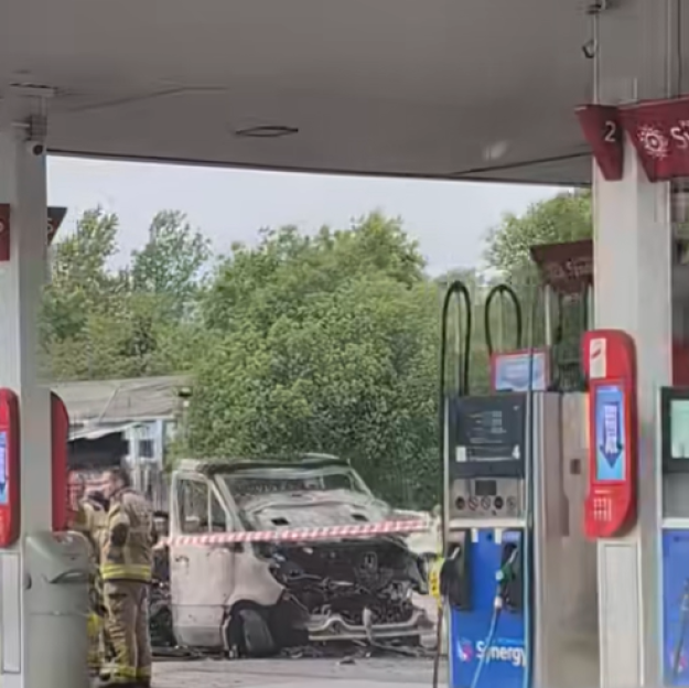 A burned campervan at a petrol station forecourt, with a firefighter standing nearby.