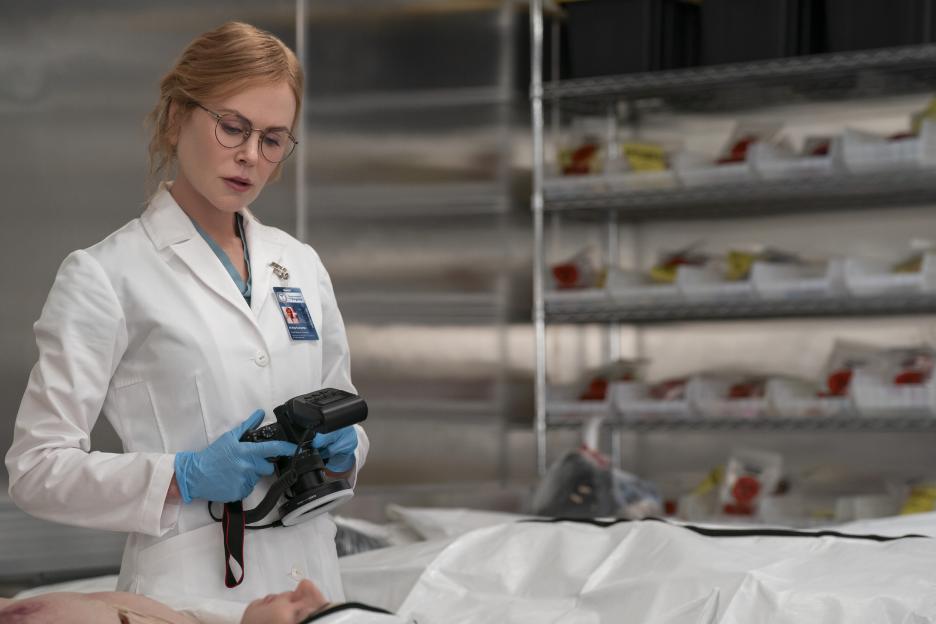 Nicole Kidman as Kay Scarpetta in a TV still, wearing glasses, a lab coat, and blue gloves, holding a camera over a body.
