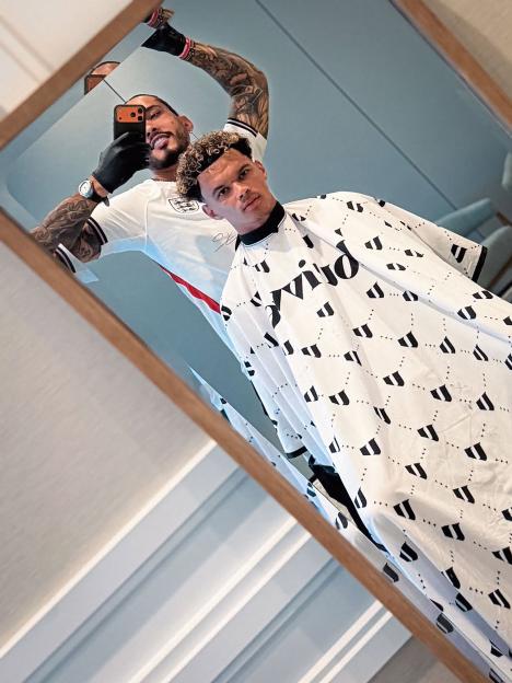 A barber in an England football shirt taking a mirror selfie with a client in a barber's cape.