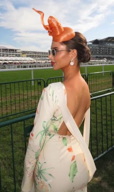 Woman wearing an orange fascinator and a floral dress at a racetrack.
