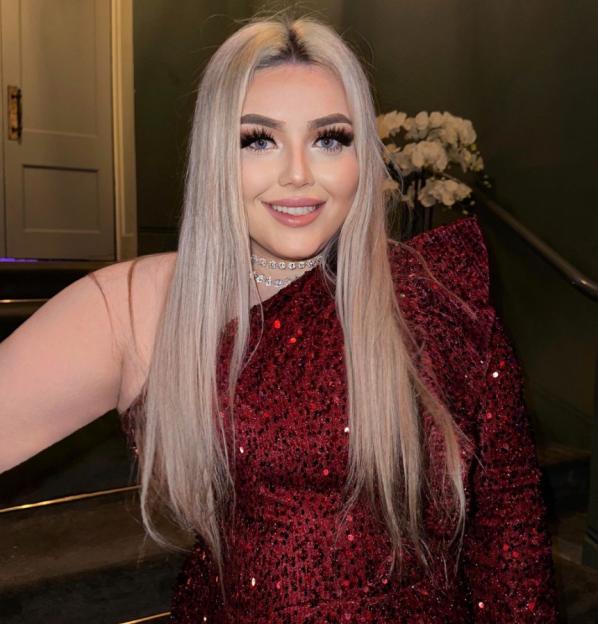 Hannah Bloor, a woman with long blonde hair, bright blue eyes, and pink eyeshadow, smiles at the camera while wearing a pink shirt.