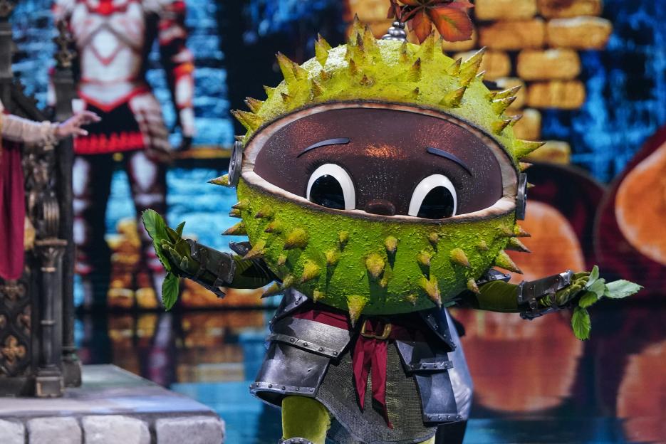 Conkers, a character from The Masked Singer, dressed in a chestnut-inspired costume with a thorny green outer shell and brown face, wearing medieval armor and holding green leaves.