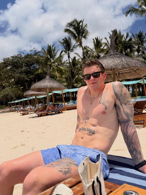 Adam Peaty on a beach wearing sunglasses and blue swimming trunks.