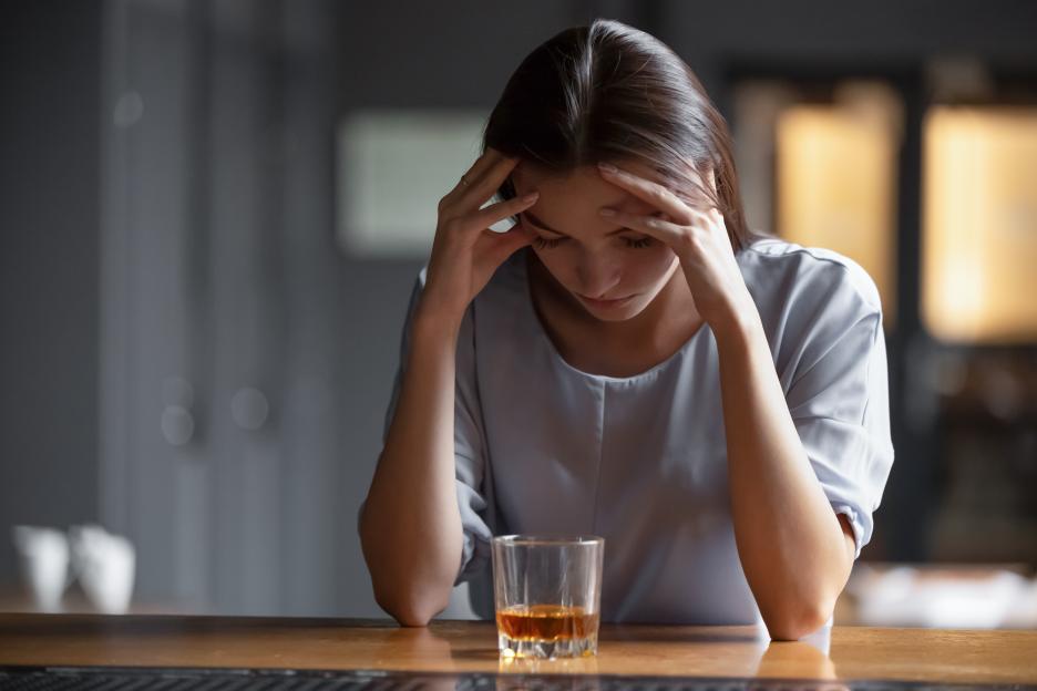 A distressed woman holding her head, with a glass of whiskey on the bar in front of her.