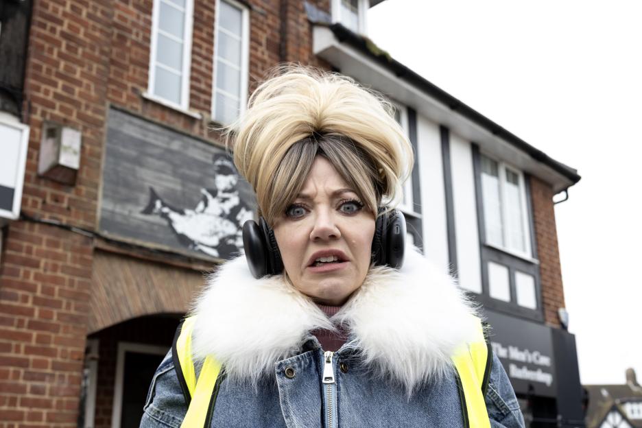 A blonde woman with headphones around her ears, a denim jacket with a white fur collar and a yellow vest looks shocked.
