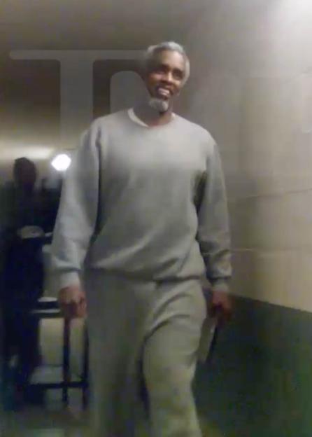 Sean "Diddy" Combs, with gray hair and beard, wearing a gray prison uniform, smiles as he walks down a hallway.