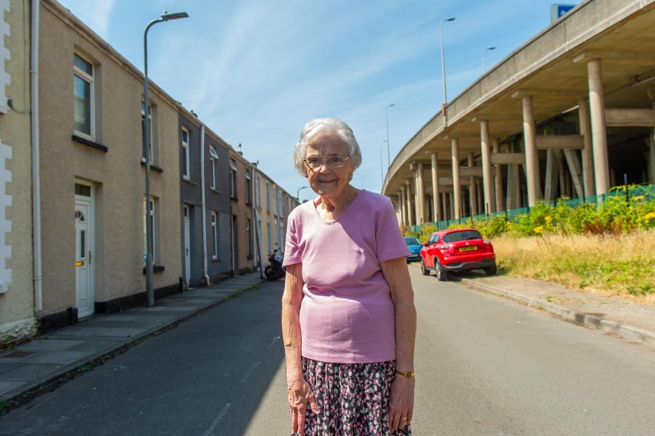 Jean Davies, 85, stands on Llewellyn Street with terraced houses to her left and the M4 motorway elevated on pillars to her right.