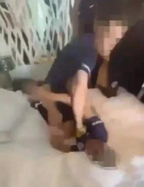 Two individuals in dark shirts are engaged in a brawl, with one person on top of another on a white bed.