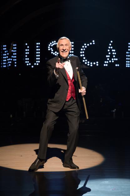 Sir Bruce Forsyth dressed in a black tuxedo with a red waistcoat, holding a golden cane, points at the viewer on a dark stage with illuminated letters in the background.