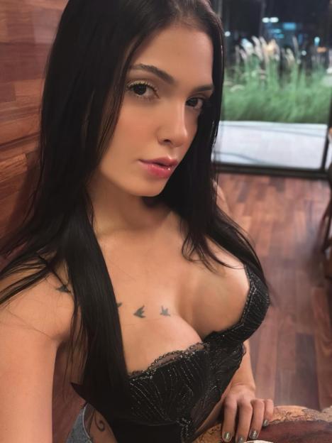 A woman with long dark hair, wearing a black top with delicate straps, looks directly at the camera.