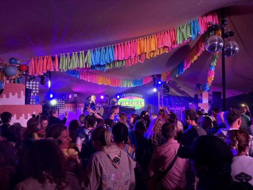 A large crowd of people dance in a tent with colorful banners, disco balls, and a stage with a DJ and a "Welcome Camp Wildfire" sign.