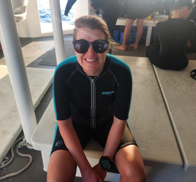 A woman in a wetsuit and sunglasses smiles on a boat with other divers.