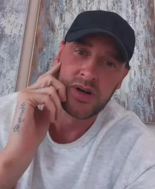 Lee Andrews wearing a black cap and white t-shirt, with a tattoo on his hand.