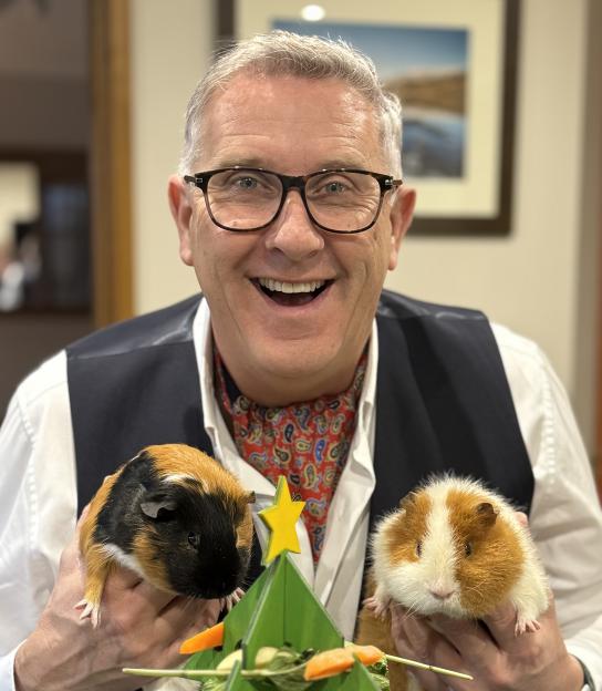 Graeme Hall smiling and holding two guinea pigs, one tricolor and one brown and white, on either side of a small green Christmas tree decoration.