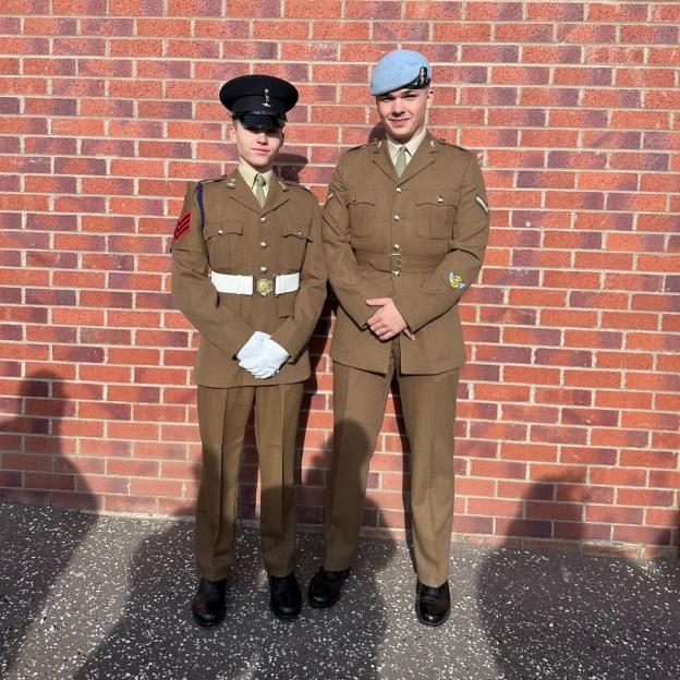 Two young men in military uniforms stand against a brick wall.