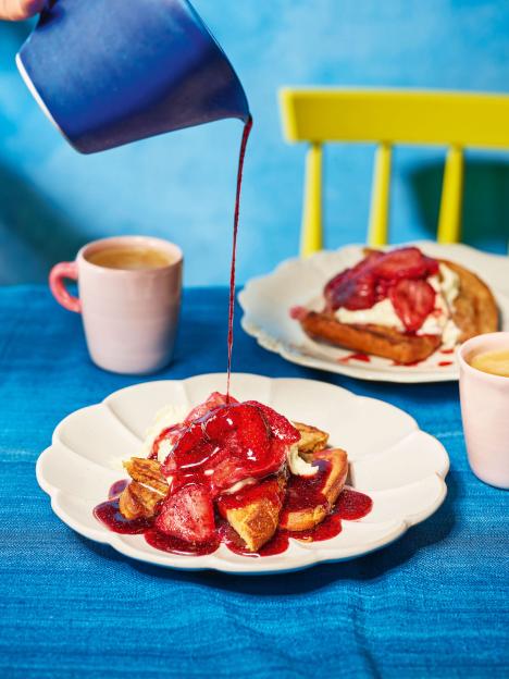 French toast with strawberries and syrup being poured over, on a blue tablecloth.