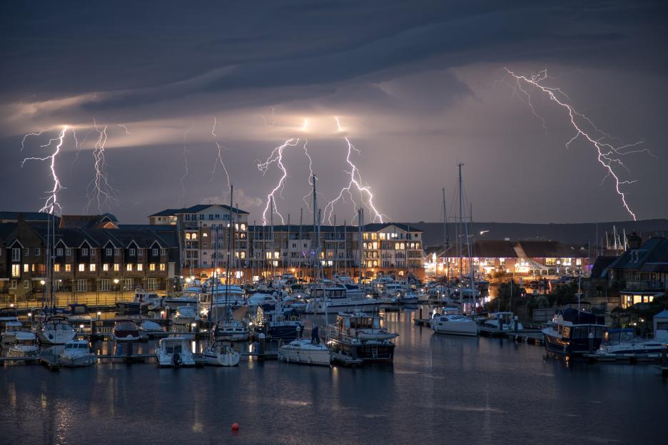 Forked lightning during a thunderstorm over Sovereign Harbour Eastbourne, with boats in the foreground and illuminated buildings in the background.