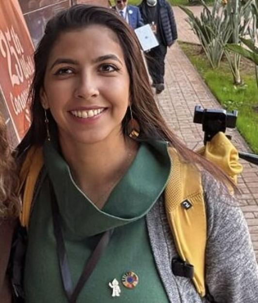 A young woman with long brown hair smiles at the camera, wearing a green cowl-neck top and a gray cardigan with a yellow backpack.