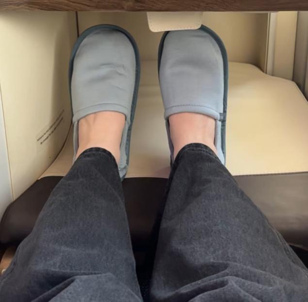 A person's feet in light blue slippers, extended into an airplane footwell.